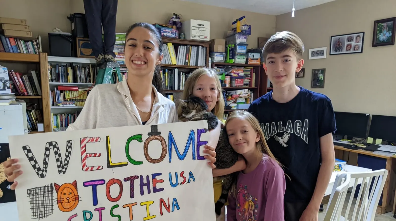 International participant stands with host siblings holding a handmade welcome sign inside an American home during a homestay experience.